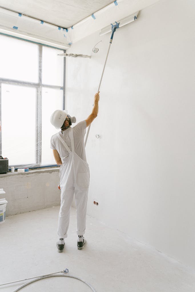 Adult painter in white uniform painting an indoor wall with a roller.